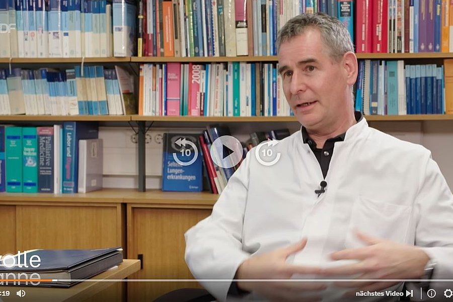 Ein Mann in einem weißen Kittel sitzt in einem Büro und gestikuliert mit beiden Händen. Im Hintergrund sind Regale mit bunten Büchern und Ordnern zu sehen.