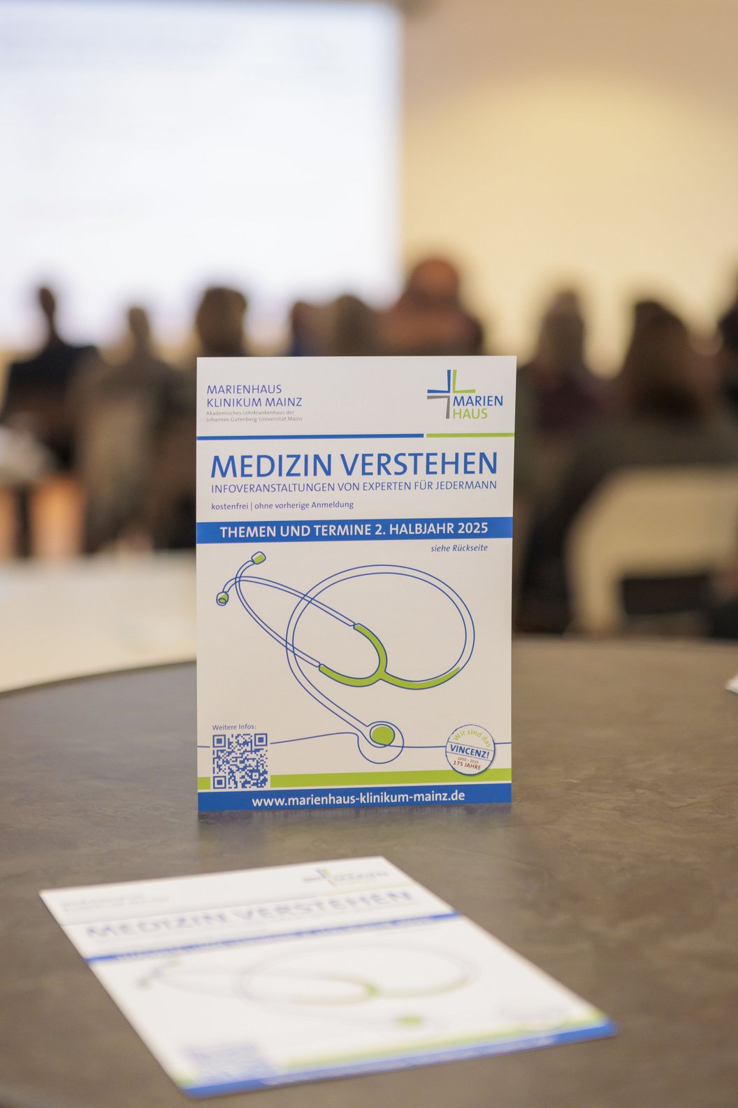Flyer mit der Überschrift 'Medizin verstehen' und einem stilisierten Stethoskop, im Hintergrund unscharfe Personen in einem Raum.