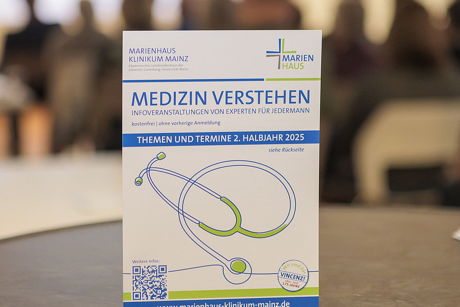 Flyer mit der Überschrift 'Medizin verstehen' und einem stilisierten Stethoskop, im Hintergrund unscharfe Personen in einem Raum.