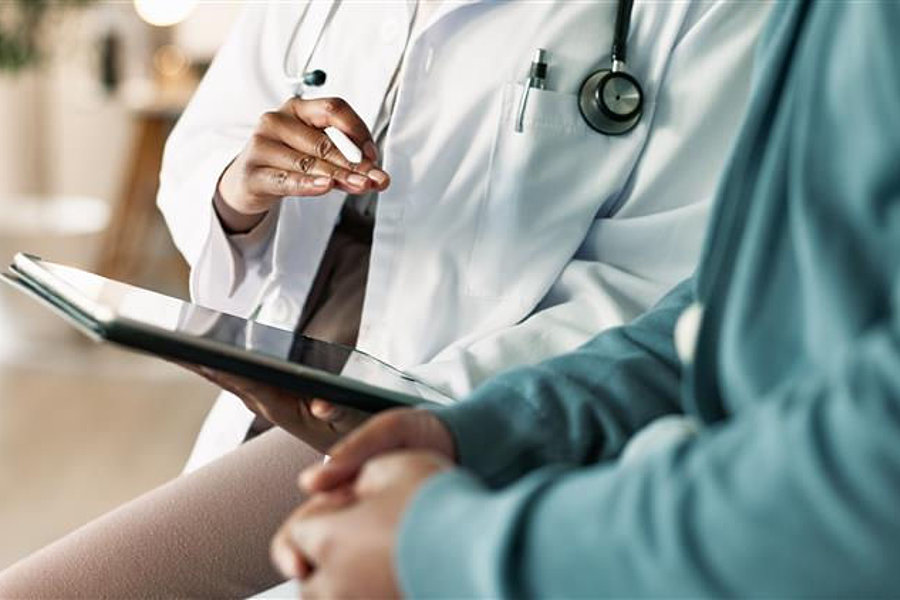 Ein Arzt in einem weißen Kittel mit einem Stethoskop sitzt neben einem Patienten. Der Arzt hält ein Tablet in der Hand und zeigt mit dem Finger darauf.
