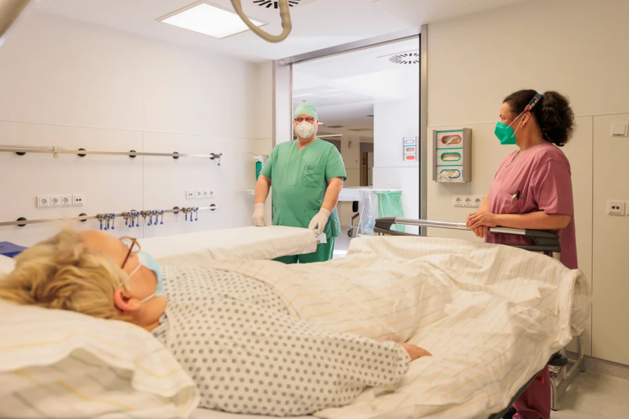 Krankenhauszimmer mit Patientin im Bett und zwei Pflegekräften in grüner und rosa Schutzkleidung