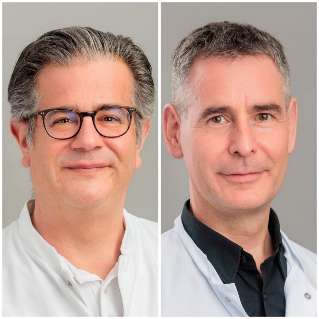 Zwei Personen in weißen Kitteln, eine mit grauen Haaren und die andere mit kurzen, dunklen Haaren. Beide stehen frontal und haben neutrale Gesichtsausdrücke.