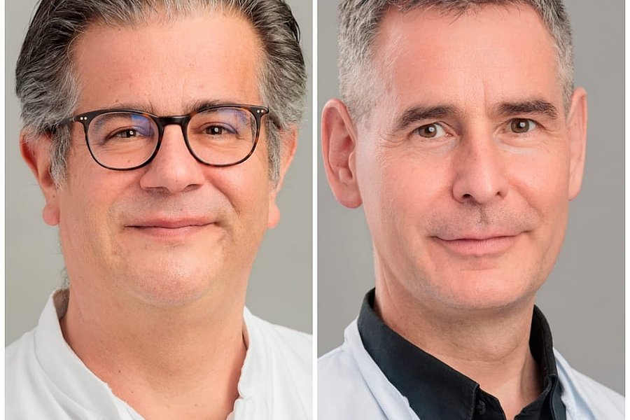 Zwei Personen in weißen Kitteln, eine mit grauen Haaren und die andere mit kurzen, dunklen Haaren. Beide stehen frontal und haben neutrale Gesichtsausdrücke.