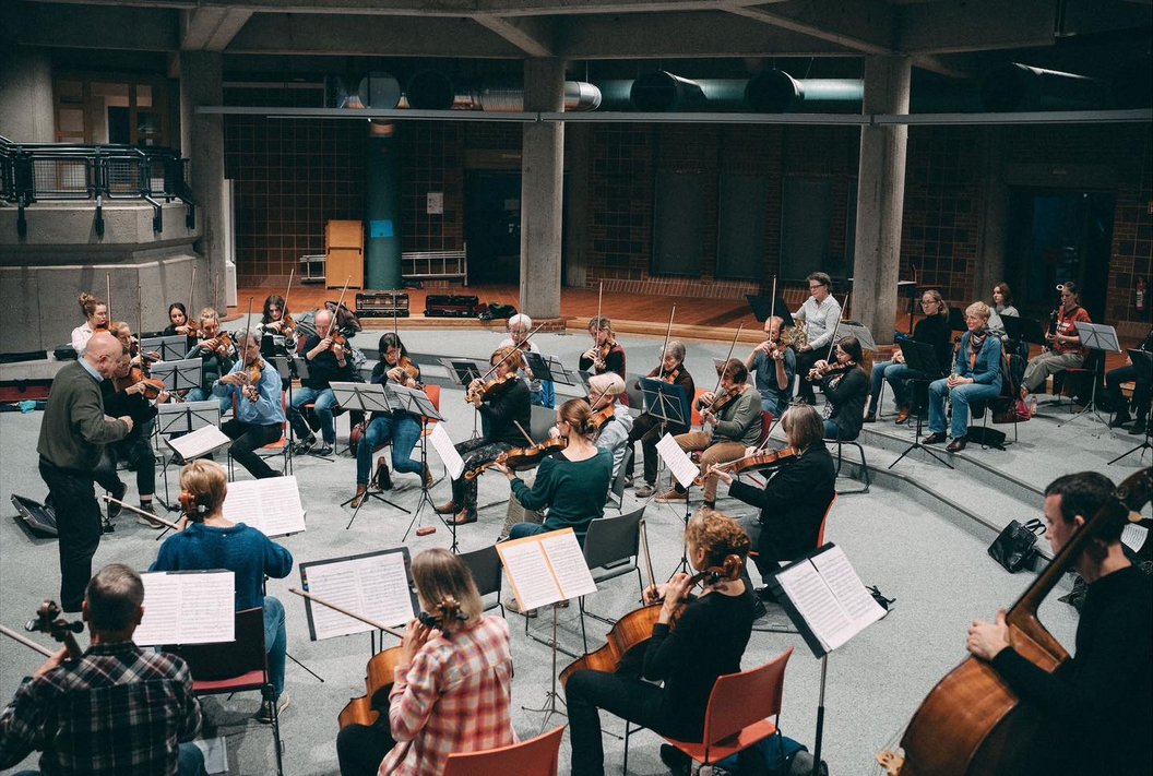 Orchesterprobe mit etwa 30 Musikern in einem großen Raum. Musiker sitzen auf Stühlen, einige mit Notenblättern vor sich. Ein Dirigent steht vorne.