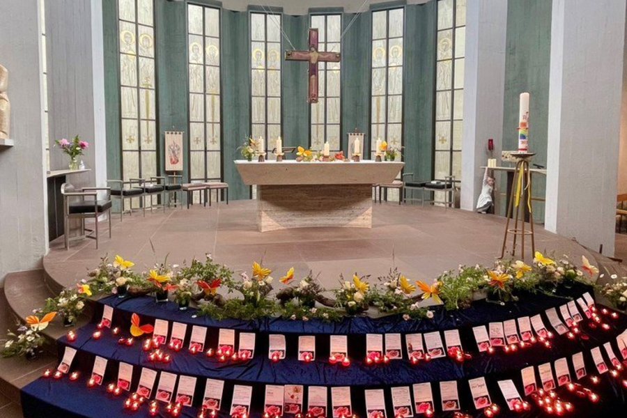 Ein runder Altar in einer Kirche, umgeben von großen Fenstern. Vor dem Altar stehen mehrere Kerzen und Blumenarrangements. Auf einer Treppe vor dem Altar liegen zahlreiche Karten mit roten Lichtern.