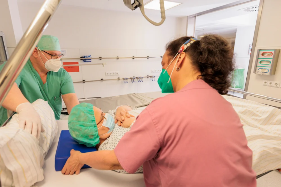 Krankenpflegerin in rosa Kleidung und grünem Mundschutz stützt Patientin mit grünem OP-Haarnetz und Maske im Krankenhausbett