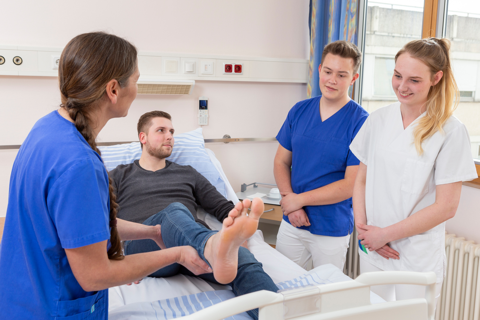 Eine Krankenschwester in blauer Uniform hält das Bein eines Patienten, der auf einem Bett liegt. Zwei weitere Personen in blauen und weißen Uniformen stehen daneben.