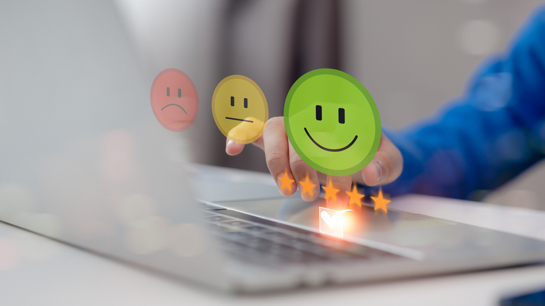 Eine Hand mit blauer Kleidung tippt auf eine Laptop-Tastatur. Über der Tastatur schweben drei emoticonartige Symbole: ein rotes trauriges Gesicht, ein gelbes neutrales Gesicht und ein grünes lächelndes Gesicht, sowie fünf goldene Sterne.