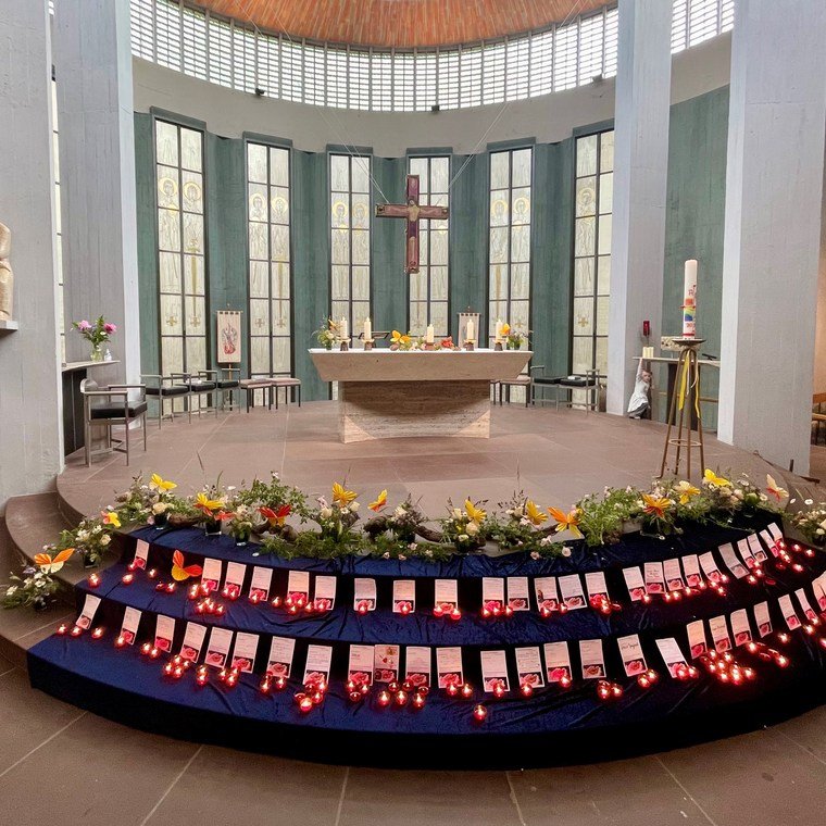 Ein runder Altar in einer Kirche, umgeben von großen Fenstern. Vor dem Altar stehen mehrere Kerzen und Blumenarrangements. Auf einer Treppe vor dem Altar liegen zahlreiche Karten mit roten Lichtern.